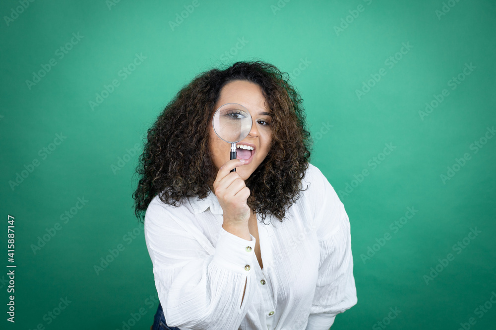Young african american girl wearing white shirt over green background surprised looking through a magnifying glass Young african american girl wearing white shirt over green background surprised looking through a magnifying glass
