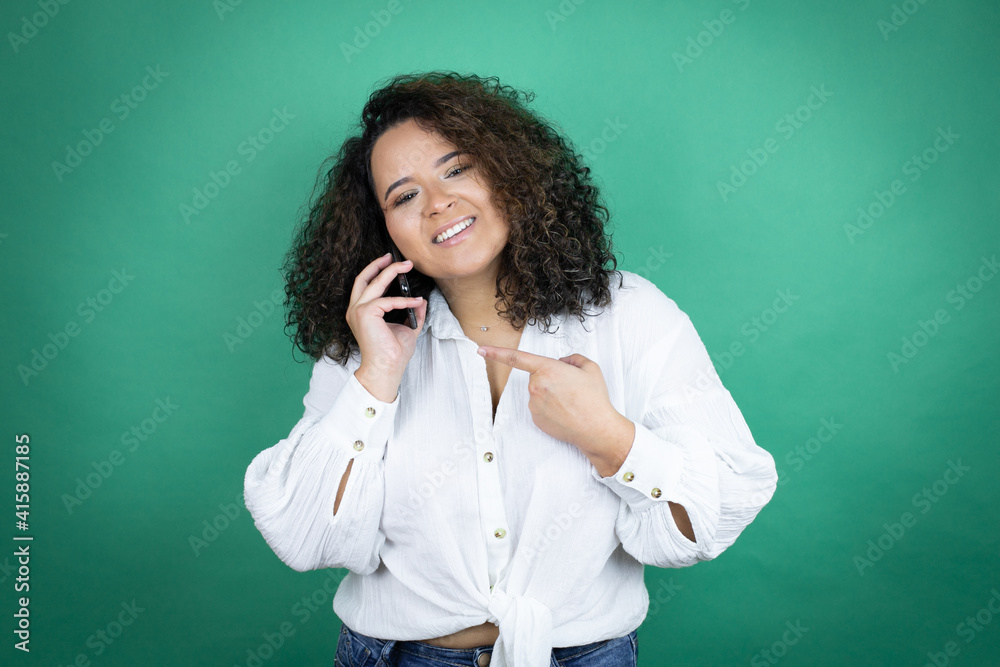 Young african american girl wearing white shirt over green background having conversation talking on the smartphone smiling happy pointing with hand and finger