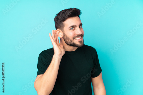Caucasian handsome man isolated on blue background listening to something by putting hand on the ear