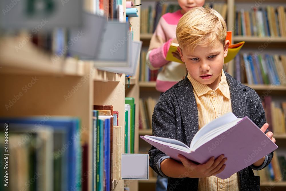 boy stands reading a book against multi colored bookshelf in library, getting knowledge boy stands reading a book against multi colored bookshelf in library, getting knowledge