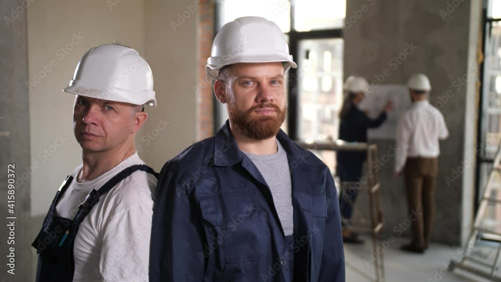 Portrait of two construction workers in helmets posing with folded arms among bare walls of apartment under renovation. Designers discussing apartment layout on blueprint blurred on background Portrait of two construction workers in helmets posing with folded arms among bare walls of apartment under renovation. Designers discussing apartment layout on blueprint blurred on background