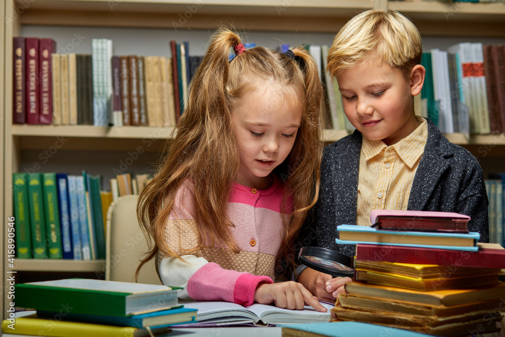 school kids reading book together while sitting at table in library, boy and girl among lots of books, preparing fro school school kids reading book together while sitting at table in library, boy and girl among lots of books, preparing fro school
