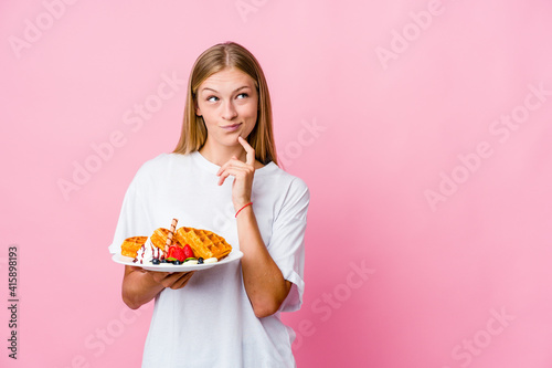 Young russian woman eating a waffle isolated making up plan in mind, setting up an idea.