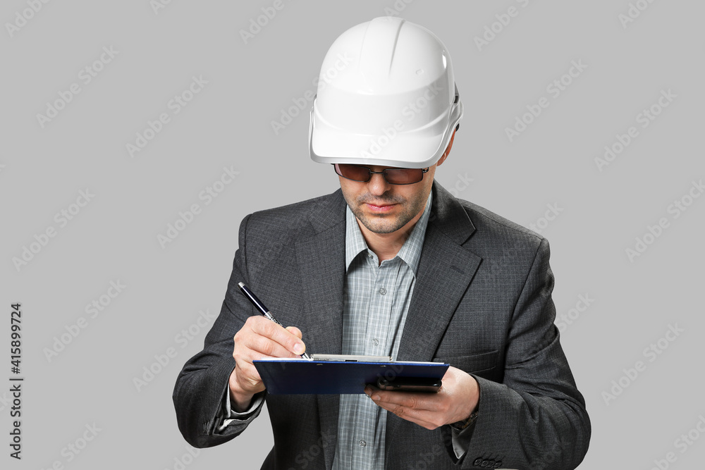 Man in suit and white safety helmet makes notes on checklist. Construction and industry, production control, isolated on gray background Man in suit and white safety helmet makes notes on checklist. Construction and industry, production control, isolated on gray background