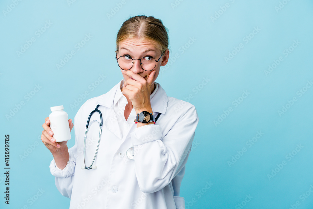 Young russian doctor woman holding pills bottle on blue thoughtful looking to a copy space covering mouth with hand. Young russian doctor woman holding pills bottle on blue thoughtful looking to a copy space covering mouth with hand.