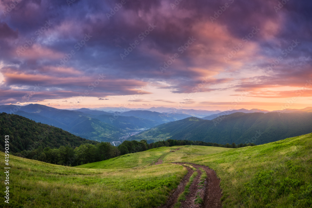 Mountain dirt road at beautiful sunset in summer. Colorful landscape with road, meadows with green grass, sky with vibrant clouds, mountains with forest. Trail on the hill. Travel and nature. Scenery