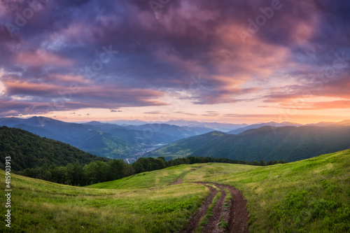 Mountain dirt road at beautiful sunset in summer. Colorful landscape with road, meadows with green grass, sky with vibrant clouds, mountains with forest. Trail on the hill. Travel and nature. Scenery © den-belitsky
