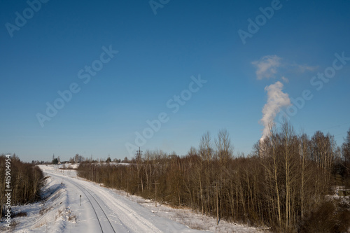 Snow-covered railway tracks between naked trees. Winter landscape. White smoke. Sunny day.