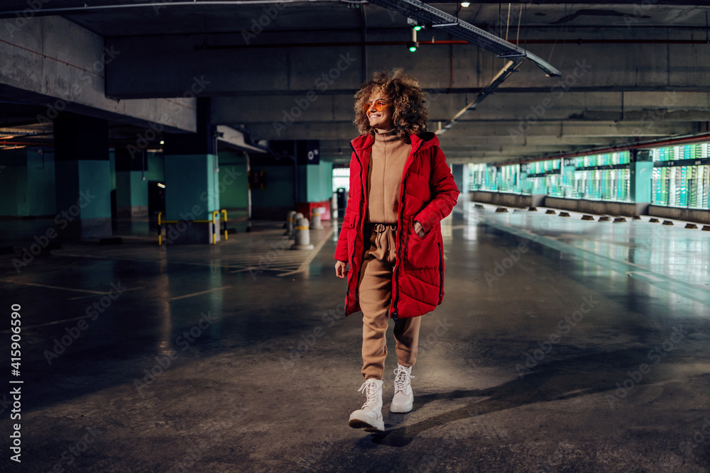 Young fashionable woman with curly hair holding hand in pocket of jacket and walking in underground garage.