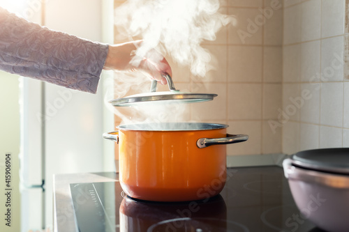 Photo Female hand open lid of enamel steel cooking pan on electric hob with boiling water or soup and scenic vapor steam backlit by warm sunlight at kitchen