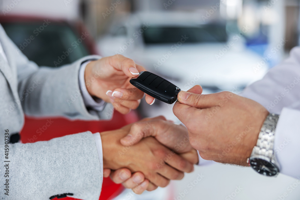 Closeup of car seller and a buyer shaking hands while standing in car salon. Seller handing car keys to a buyer.