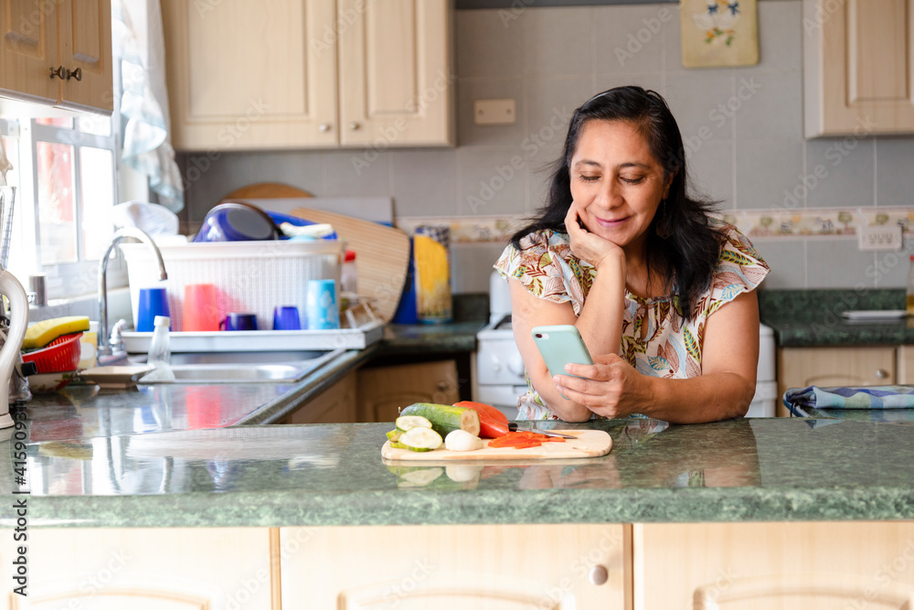 Hispanic mom searching recipes on her phone-woman preparing healthy and organic salad while checking her cell phone-housewife cooking while she looks at a phone