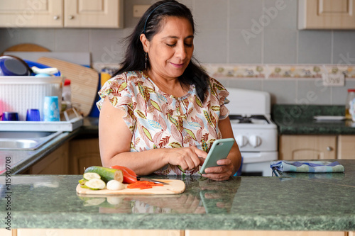 Hispanic mom searching recipes on her phone-mature woman preparing healthy and organic salad while checking her cell phone-housewife cooking while she looks at a phone