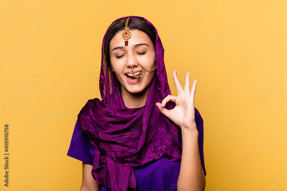 Young Indian woman wearing a traditional sari clothes isolated on yellow background winks an eye and holds an okay gesture with hand. Young Indian woman wearing a traditional sari clothes isolated on yellow background winks an eye and holds an okay gesture with hand.