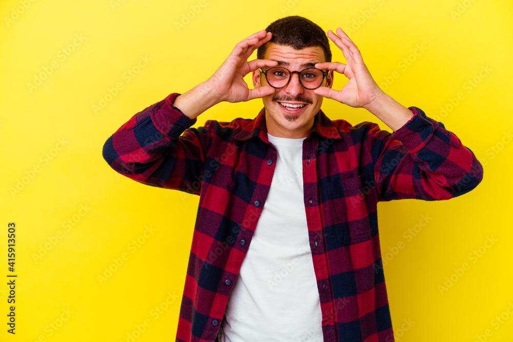 Young caucasian cool man isolated on yellow background showing okay sign over eyes Young caucasian cool man isolated on yellow background showing okay sign over eyes