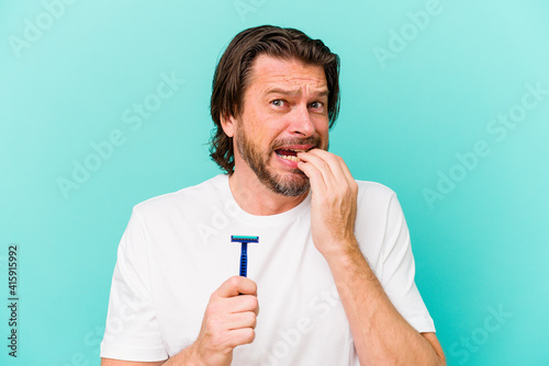 Middle age dutch man holding a razor blade isolated on blue background biting fingernails, nervous and very anxious.