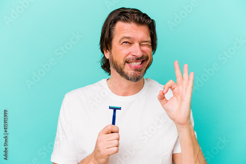 Middle age dutch man holding a razor blade isolated on blue background cheerful and confident showing ok gesture.