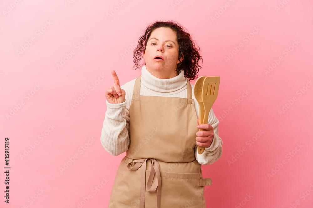 Woman with Down syndrome cooking at home isolated on pink background pointing upside with opened mouth. Woman with Down syndrome cooking at home isolated on pink background pointing upside with opened mouth.