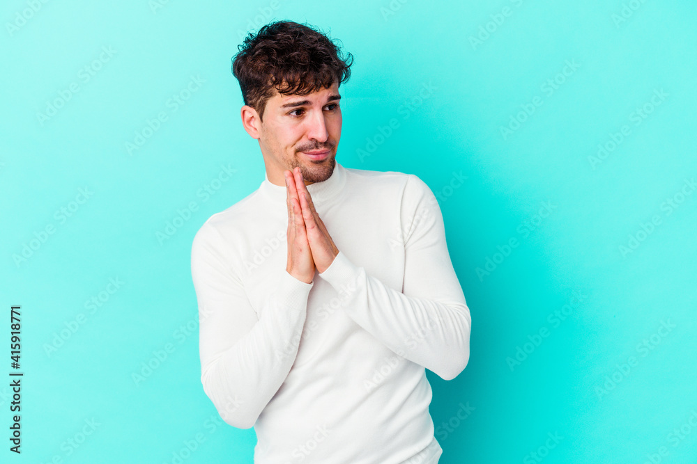 Young caucasian man isolated on blue background praying, showing devotion, religious person looking for divine inspiration. Young caucasian man isolated on blue background praying, showing devotion, religious person looking for divine inspiration.