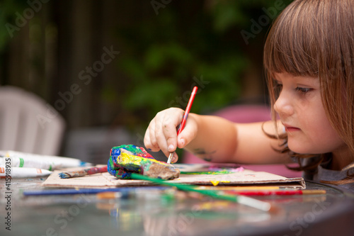 Young Girl paints Rocks with colorful Paints and Brushes outside to be creative outside on a sunny summer day and surrounded by nature