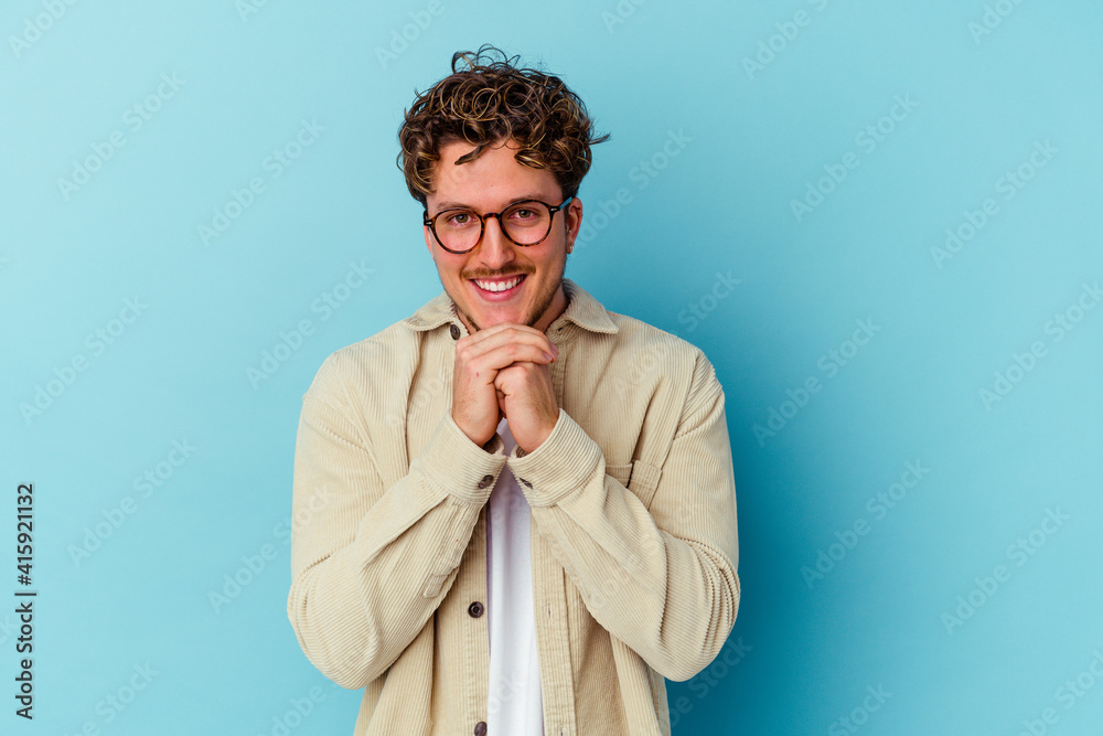 Young caucasian man wearing eyeglasses isolated on blue background keeps hands under chin, is looking happily aside. Young caucasian man wearing eyeglasses isolated on blue background keeps hands under chin, is looking happily aside.