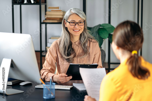 Friendly female mature hr manager listening and looks a resume of a new applicant introduction for a job. Gray-haired female employer talks and meets with female job seeker. photo