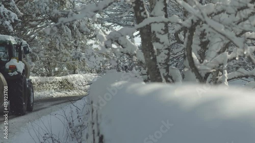 Tractor passing slowly on snowy country tree lined road in daytime mid winter. photo