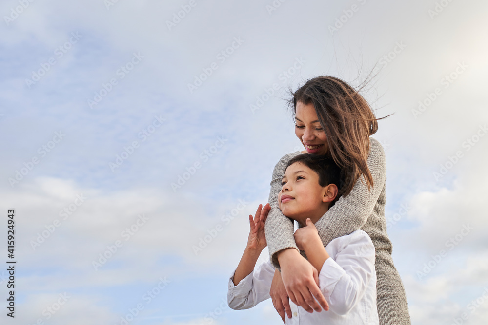 Young mother hugging her child, blue sky background with clouds. Copy space. Young mother hugging her child, blue sky background with clouds. Copy space.