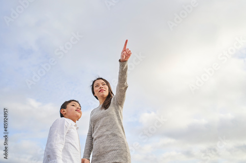 Young Latina mother with child pointing to some point against a bright blue sky with clouds. Family concept.