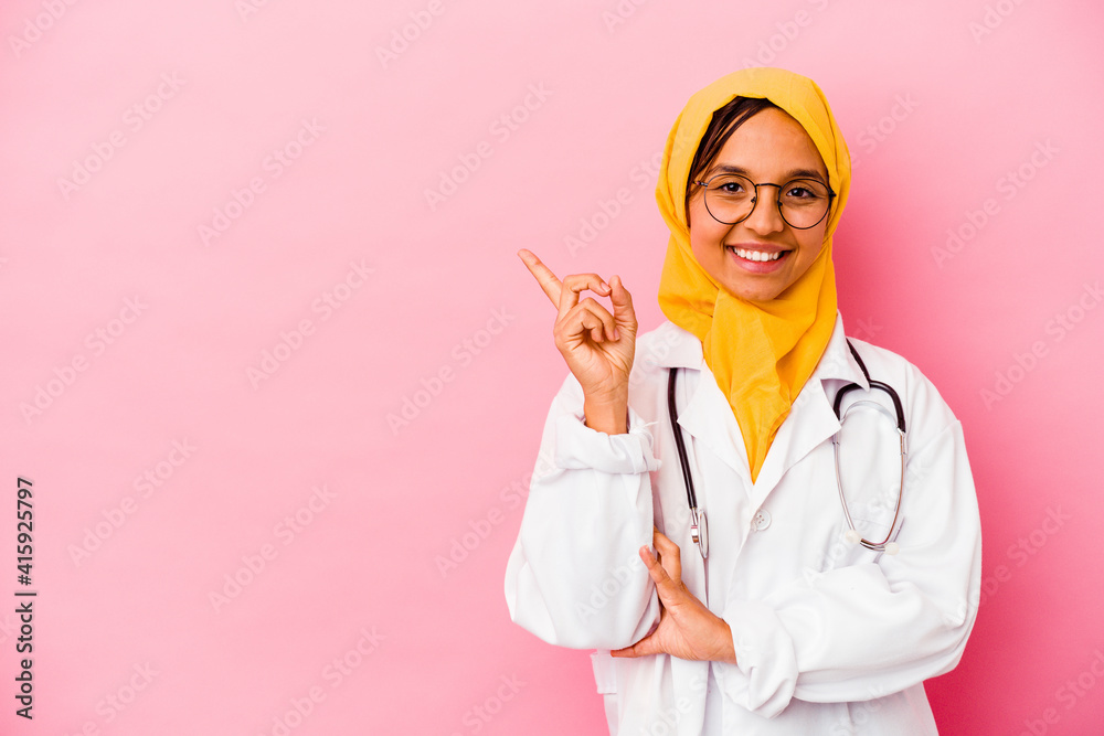 Young doctor muslim woman isolated on pink background smiling cheerfully pointing with forefinger away. Young doctor muslim woman isolated on pink background smiling cheerfully pointing with forefinger away.