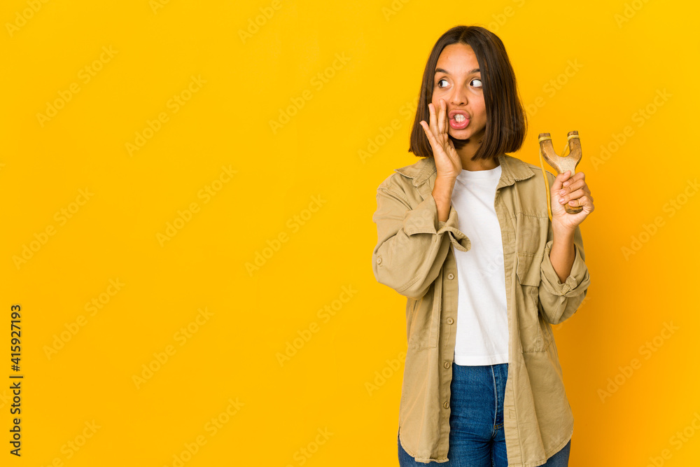 Young hispanic woman holding a slingshot is saying a secret hot braking news and looking aside Young hispanic woman holding a slingshot is saying a secret hot braking news and looking aside