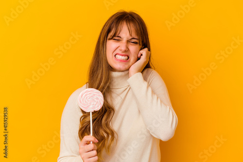 Young caucasian woman holding a lollipop isolated on yellow background covering ears with hands.