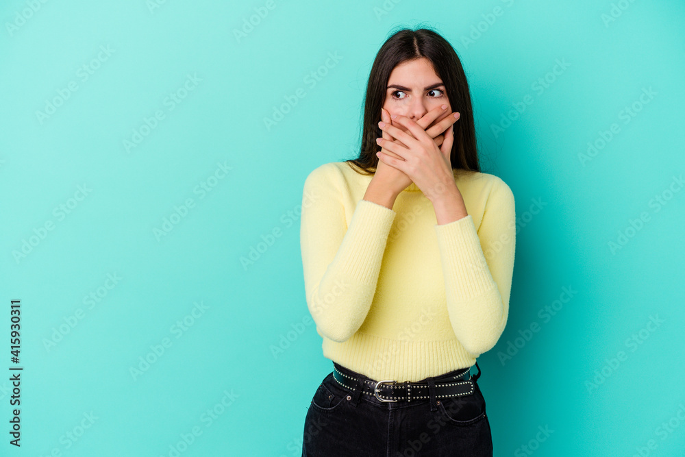 Young caucasian woman isolated on blue background thoughtful looking to a copy space covering mouth with hand. Young caucasian woman isolated on blue background thoughtful looking to a copy space covering mouth with hand.