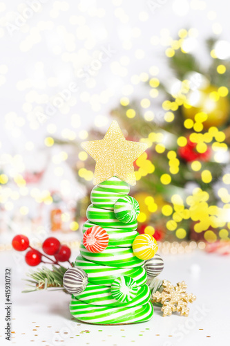 Caramel candy Christmas tree. Christmas card concept. Striped christmas caramel tree with golden star on top. Light up natural green fir tree with bokeh in background. Candle  holly berries  confetti.