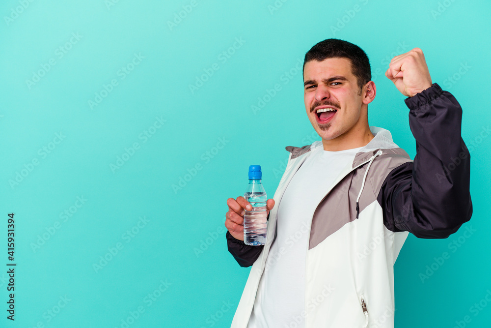 Young sporty caucasian man drinking water isolated on blue background raising fist after a victory, winner concept. Young sporty caucasian man drinking water isolated on blue background raising fist after a victory, winner concept.