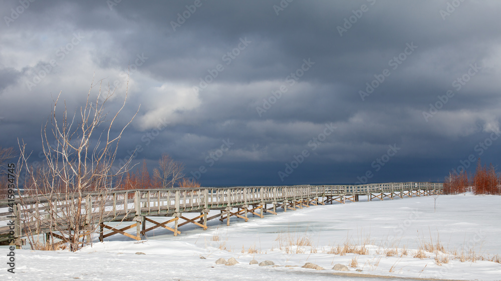 An old, wooden bridge crosses a section of frozen Georgian Bay waterfront in Collingwood, Ontario. The sky above is menacing and gloomy, with a winter storm approaching. Empty walking trail. An old, wooden bridge crosses a section of frozen Georgian Bay waterfront in Collingwood, Ontario. The sky above is menacing and gloomy, with a winter storm approaching. Empty walking trail.