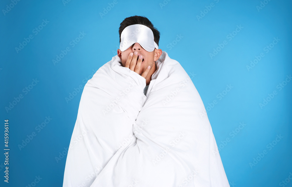 Man wrapped with blanket in sleeping mask yawning on blue background Man wrapped with blanket in sleeping mask yawning on blue background