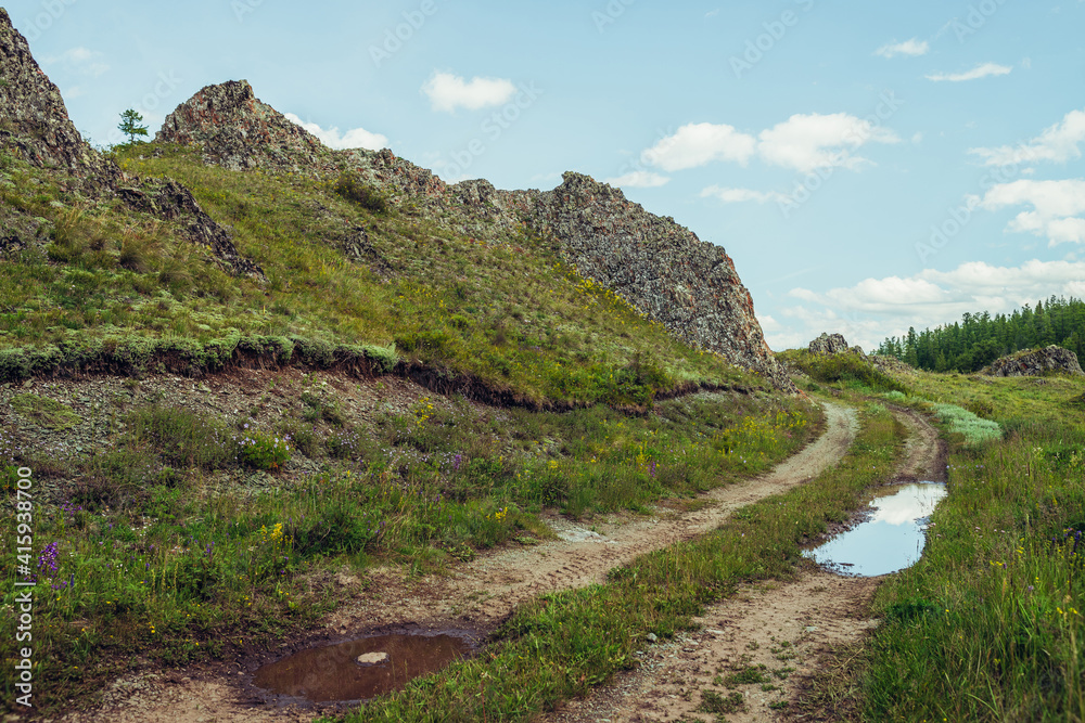 Scenic alpine landscape with dirt road along rocks in highlands. Puddle on road near rocks and lush vegetation in mountains. Beautiful mountain scenery with dirt road with puddle among mountain flora.