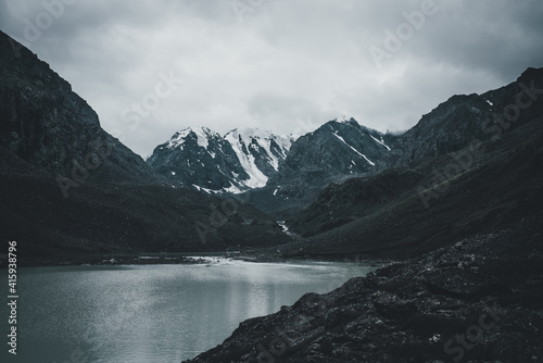 Dark atmospheric landscape with mountain lake among deep black rocks on background of snowy mountains under gray cloudy sky. Bleak view to mountain lake and glacier in deep black colors. Dark tones.
