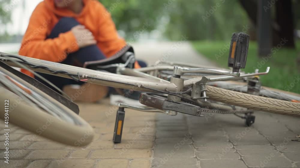 Close-up of bicycle lying on park alley with blurred young woman rubbing injured knee at background. Millennial Caucasian lady fell from bike on summer day. Sport lifestyle.