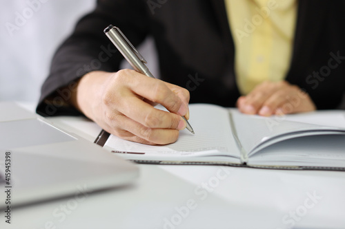 Businesswoman hands in black suit sitting and writing notebook and working or using computer on white table with business chart and calculator in modern office with paper work