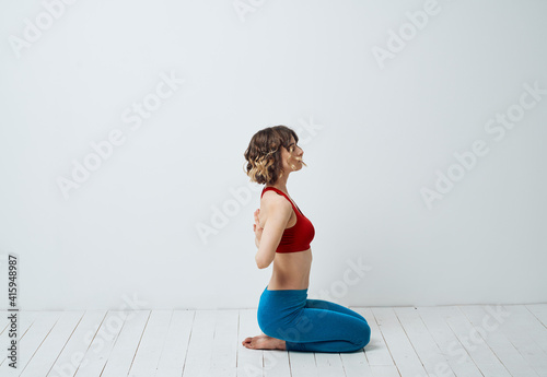 A woman in blue leggings sits on a light floor indoors and holds her hands behind her back