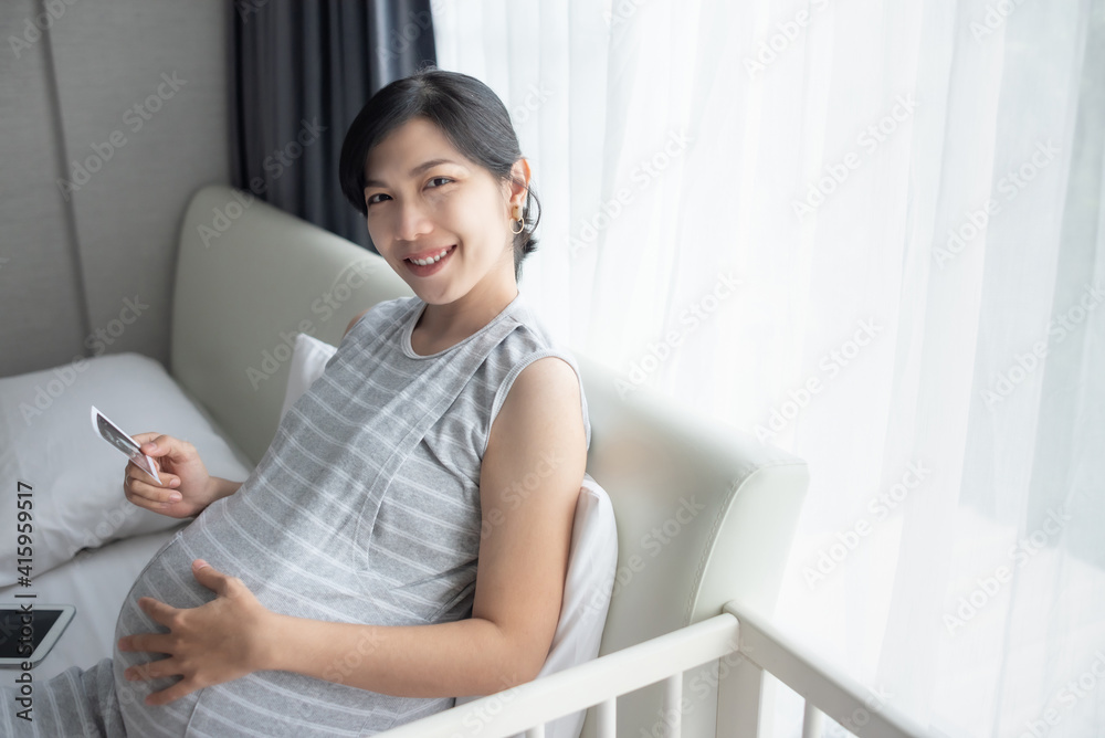 Happy Smiling Asian Pregnant woman sitting on bed touching her belly and holding ultrasound scan photo. Mother with sonogram of her unborn baby. Concept of pregnancy, Maternity prenatal care Happy Smiling Asian Pregnant woman sitting on bed touching her belly and holding ultrasound scan photo. Mother with sonogram of her unborn baby. Concept of pregnancy, Maternity prenatal care