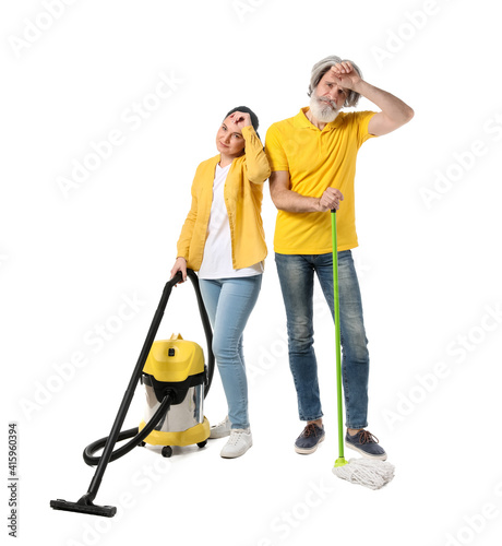 Tired mature couple with vacuum cleaner and floor mop on white background