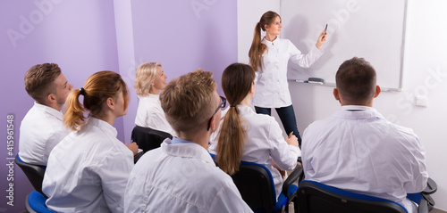 Female medical student lecturing near whiteboard in front of teacher and group of students in auditorium.
