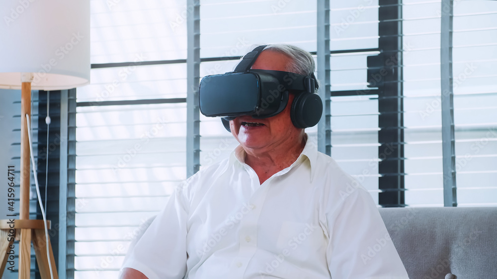 Asian senior man having fun playing video game with virtual reality glasses at home.
