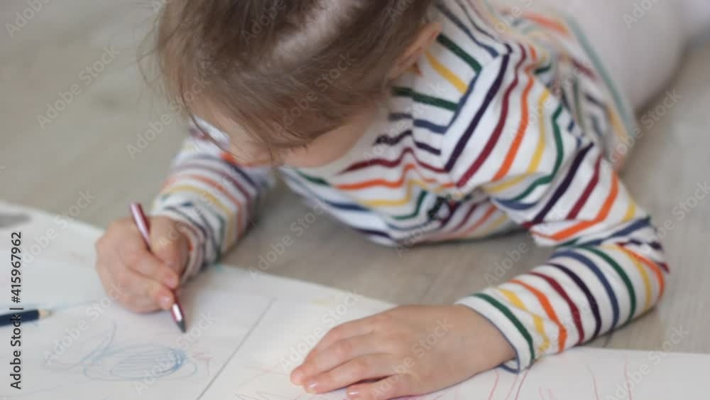 a little girl in a colorful T-shirt lies on the floor and draws drawings in an album with a colored pencil a little girl in a colorful T-shirt lies on the floor and draws drawings in an album with a colored pencil