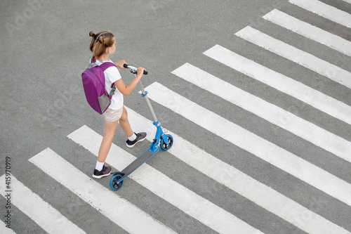 Schoolgirl carrying scooter and crossing road on way to school. Zebra traffic walk way in the city. Pedestrian passing a crosswalk. Stylish kid walking with backpack. Safety concept for road users