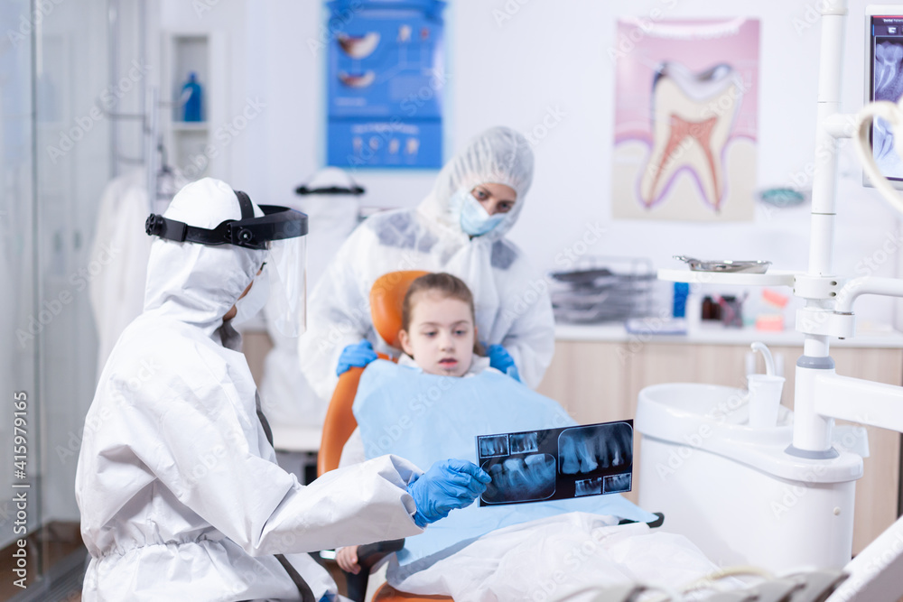Dentist dressed in ppe suit reviewing panoramic mouth radiography of little girl patient. Stomatolog in protectie suit for coroanvirus as safety precaution holding child teeth x-ray during Dentist dressed in ppe suit reviewing panoramic mouth radiography of little girl patient. Stomatolog in protectie suit for coroanvirus as safety precaution holding child teeth x-ray during