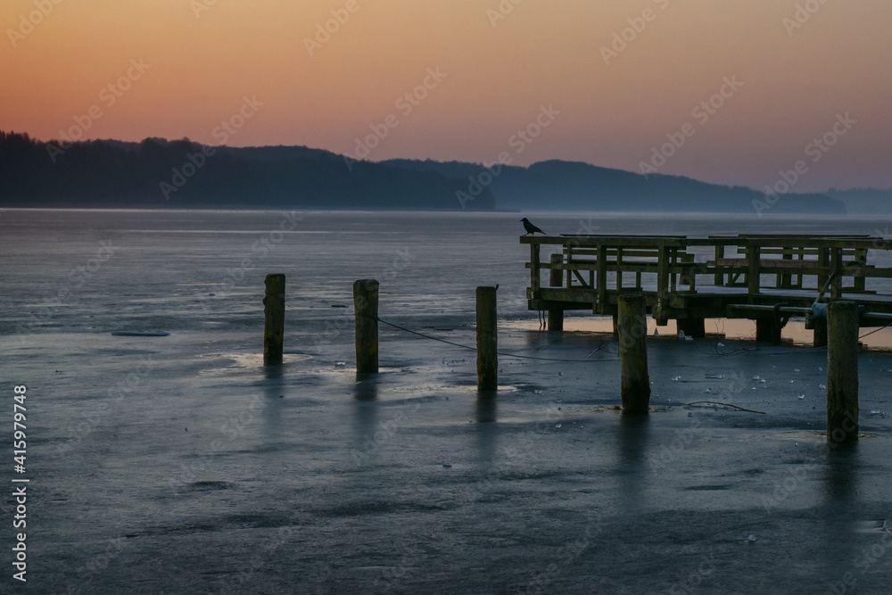 Dawn over a frozen lake and a wooden jetty with a crow enjoying the cold morning landscape, copy space